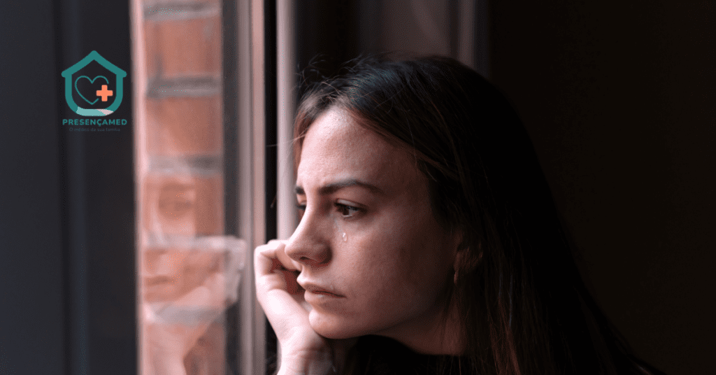 Mulher jovem sentada sozinha olhando pela janela em dia nublado, com expressão pensativa, representando os sintomas de depressão e isolamento emocional